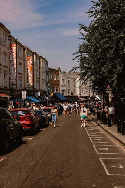 A busy street scene in Earls Court showing narrow pavement with parked cars along the side, and a row of multi-storey buildings with retail shops on the ground floor. Pedestrians, some carrying boxes or bags, walk along the sidewalk, while others stand or chat near the shop entrances. In the background, a white van from Movers Earls Court is partially visible as it loads or unloads household furniture and packing materials, including cardboard boxes, fabric-wrapped items, and plastic wrap, onto the pavement close to the storefronts. The scene is outdoors under a partly cloudy sky; tall trees provide shade over parts of the street, and a black bollard indicates a designated parking or loading zone, supporting the context of furniture transport and home relocation within a city environment.