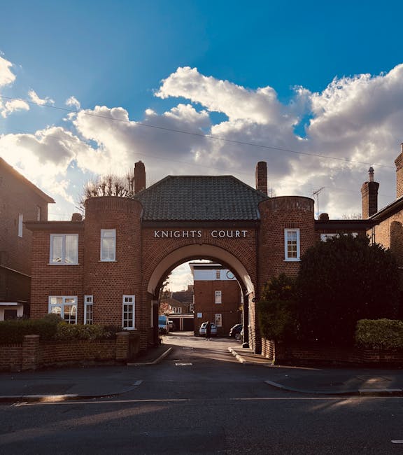 A brick archway labeled 'Knights Court' serves as the entrance to a residential area, with two symmetrical buildings on either side featuring white-framed windows and small bushes in front. The archway leads to a courtyard with parked cars and a narrow street beyond, under a partly cloudy sky with bright sunlight illuminating the scene. This image reflects a typical suburban setting in Earls Court, where home relocation and furniture transport often involve navigating narrow streets and parking restrictions. Movers Earls Court may use this location for loading or unloading household items during a house removal service, with equipment such as trolleys and blankets potentially seen nearby for the safe transfer of furniture and boxes. The scene emphasizes the importance of careful planning for moving logistics in areas with limited space.
