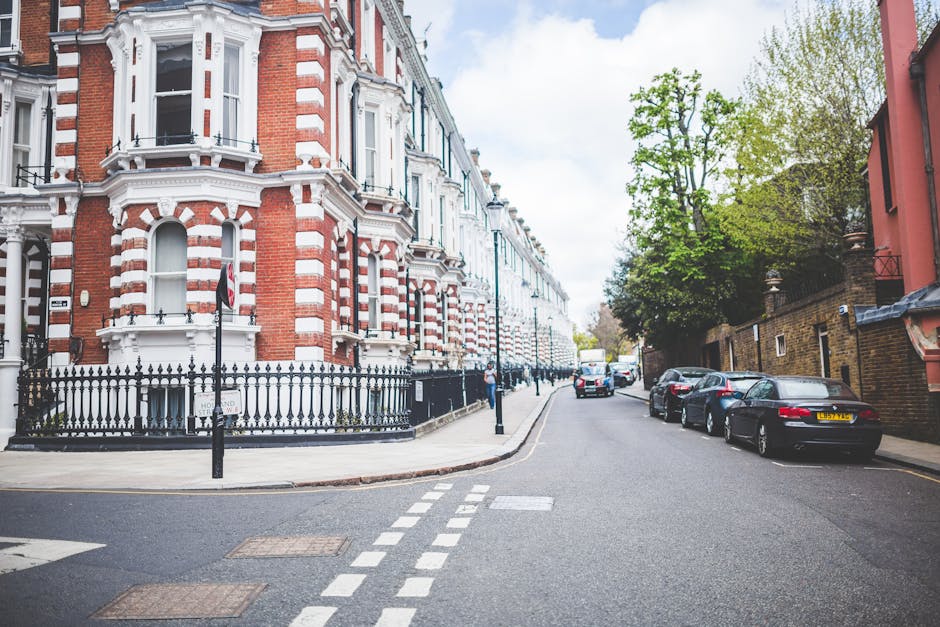 A busy street scene in Earls Court showing narrow pavement with parked cars along the side, and a row of multi-storey buildings with retail shops on the ground floor. Pedestrians, some carrying boxes or bags, walk along the sidewalk, while others stand or chat near the shop entrances. In the background, a white van from Movers Earls Court is partially visible as it loads or unloads household furniture and packing materials, including cardboard boxes, fabric-wrapped items, and plastic wrap, onto the pavement close to the storefronts. The scene is outdoors under a partly cloudy sky; tall trees provide shade over parts of the street, and a black bollard indicates a designated parking or loading zone, supporting the context of furniture transport and home relocation within a city environment.