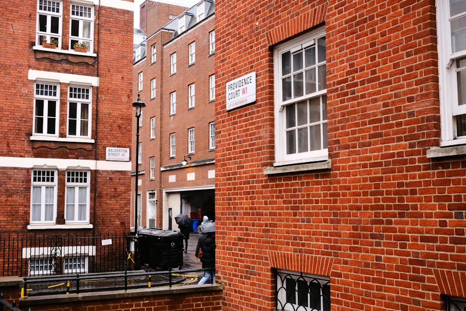 A view of a residential street in Earls Court with red brick buildings and white-framed windows. In the foreground, a section of a brick building has a window with white muntins, and a metal security grille on a lower window. On the narrow street, two movers from Movers Earls Court, dressed in dark clothing and caps, are loading large cardboard boxes and wrapped pieces of furniture into a black moving van parked at the curb. The van is positioned next to an alleyway where additional boxes and packing materials are visible, along with a small black waste bin. A street sign attached to a lamppost indicates the intersection of Balderton Street and Providence Court W1, while another sign on the building reads 'Providence Court W1.' The scene is illuminated by natural daylight, illustrating the process of packing and furniture transport during a home relocation, aligning with the topic of avoiding hidden charges on Earls Court removals.