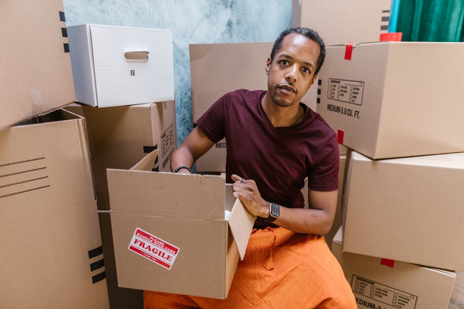 A man with dark hair and a serious expression is inside a room surrounded by stacked cardboard boxes of various sizes, some sealed with packing tape, and one labeled 'Fragile.' He is wearing a maroon T-shirt, orange pants, and a smartwatch, and is holding an open cardboard box in his lap while looking towards the camera. The environment appears to be an indoor space prepared for a home relocation, with boxes arranged for packing or unpacking. Visible in the background are a blue wall, a partially visible doorway, and additional packed boxes, indicating the process of packing or furniture transport typical in house removals. The scene reflects activities involved in furniture transport and packing during a move, with the presence of packing materials, such as cardboard and tape, and equipment like boxes and possibly moving blankets or straps implied to aid in safe loading and unloading. This visual supports content on how to avoid hidden charges on Earls Court removals, as provided by Movers Earls Court, by illustrating the packing and loading process involved in professional removals.